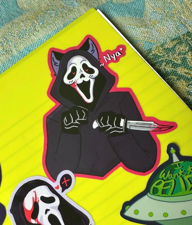 May include: A sticker of a black-hooded figure with a white mask and cat ears holding a bloody knife. The figure has red eyes and a wide, open mouth. The sticker has a pink border and the text "~Nyaa" is written in pink above the figure.
