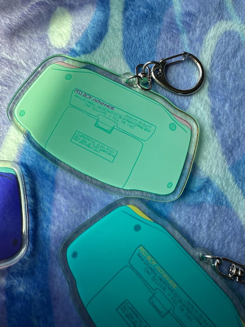 May include: A green and yellow Game Boy Advance keychain with the text "PISS BOY ADVANCE" and "MAN IN CHHEUS CAN TREZMAS FST IN URMUTH PUVAD BU CANT BELIEVE U FELL FOR THAT" printed on it.