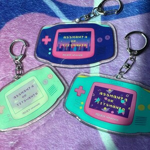 May include: Three keychain charms shaped like a handheld video game console. Each charm is a different color: blue, green, and pink. The screen on each charm displays the text "ASSMAN? A OR TITSNAN? B" and the text "PISS BOY ADVANCE".
