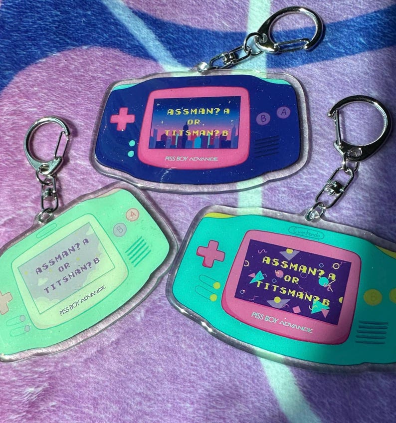 May include: Three keychain charms shaped like a handheld video game console. Each charm is a different color: blue, green, and pink. The screen on each charm displays the text "ASSMAN? A OR TITSNAN? B" and the text "PISS BOY ADVANCE".