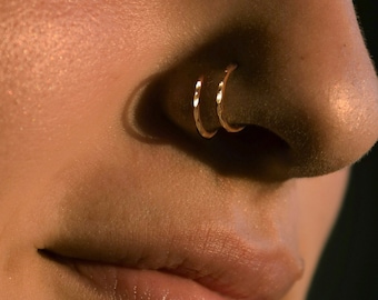 May include: Close-up of a person's nose with two gold nose rings. The ring closer to the nose tip is a pierced hoop, while the ring closer to the cheek is a faux hoop. The text "Right twist in Right nostril 18 gauge, 10mm Pierced hoop Right closer to nose tip Faux hoop Left closer to cheek" is visible below the image.