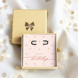 May include: A small pink gift box with a gold lid. The box has a pink and white pattern with the words "Happy Birthday" on the front. The box is open and shows a pair of small silver earrings inside.