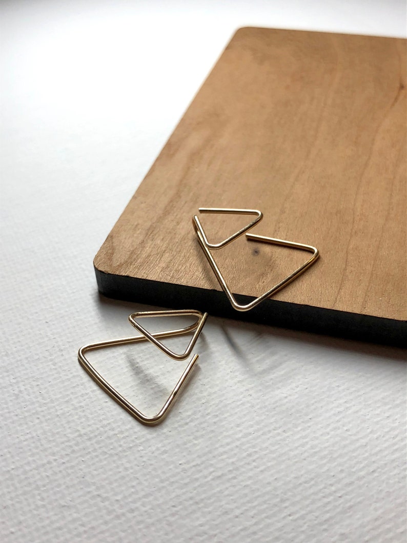 K&ouml;nnte beinhalten: Goldene Dreiecksohrringe mit einem mehrschichtigen Design. Die Ohrringe liegen auf einer Holzoberfl&auml;che.