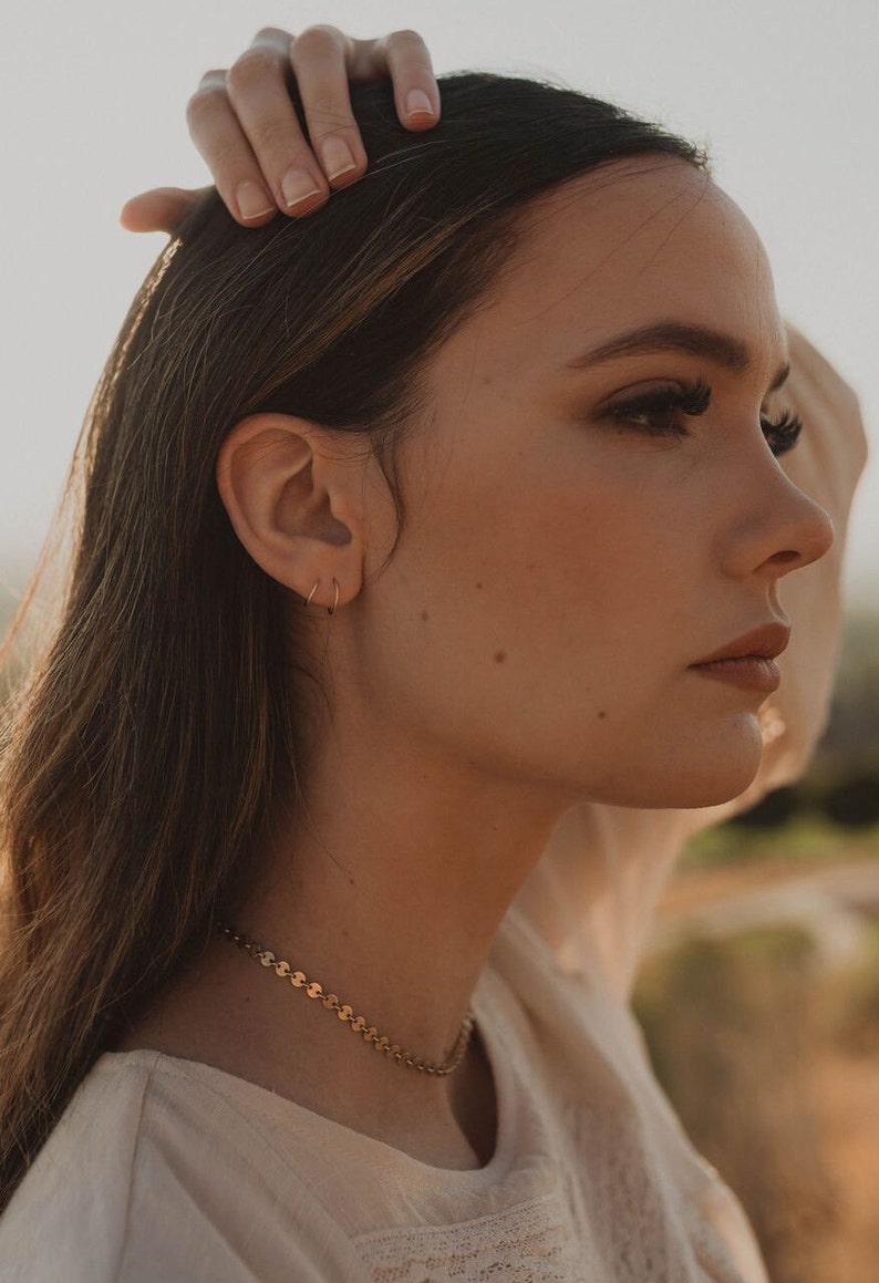 May include: A woman with long brown hair wearing a gold chain necklace and small gold hoop earrings. She is wearing a white blouse with a delicate lace pattern.