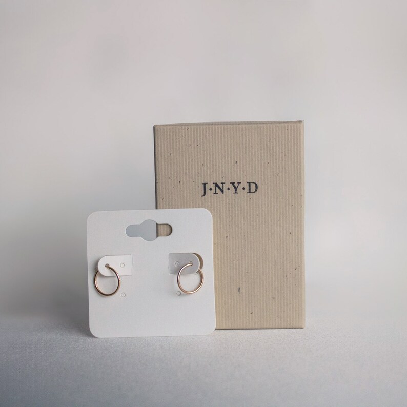 K&ouml;nnte beinhalten: Ein Paar goldene Creolen auf einer wei&szlig;en Karte mit dem Markennamen "J.N.Y.D." auf einem braunen Karton.