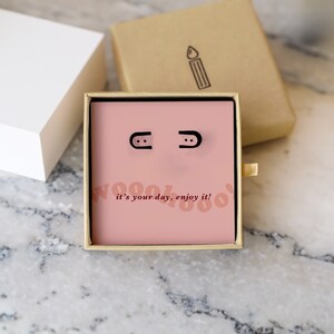 May include: A pink box with a black and white graphic that says "Woohoo! It's your day, enjoy it!"  The box contains a pair of black stud earrings.
