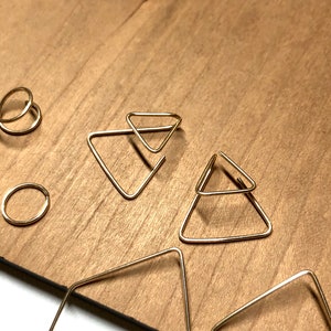 K&ouml;nnte beinhalten: Ein Set goldener geometrischer Ohrringe mit einem Paar Doppel-Dreiecks-Ohrringen und einem Paar quadratischen Creolen.