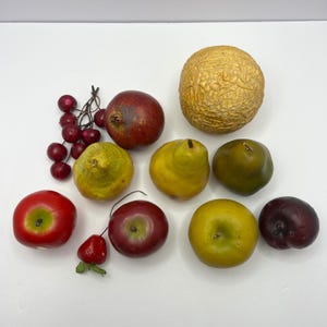 May include: A collection of artificial fruit including a cluster of dark red cherries, a deep red pomegranate, a textured gold melon, two yellow-green pears, a dark green pear, a bright red apple, a dark red apple, a yellow apple, and a plum.