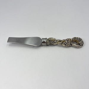 May include: A silver-toned metal cheese spreader with a flat, smooth blade and a decorative handle featuring a detailed, raised relief pattern of grape clusters and vine leaves. The handle has a metallic finish with darkened recessed areas.