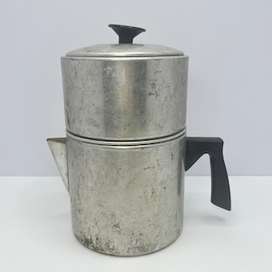 May include: A silver-toned metal coffee percolator with a weathered, textured surface. The cylindrical vessel features a black plastic handle on the side, a small triangular spout, and a matching black knob on the removable lid at the top.