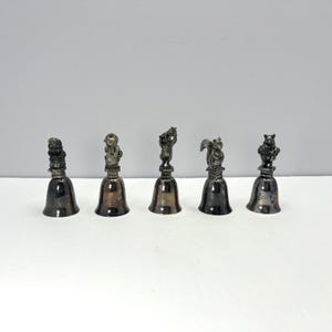 May include: A set of five silver-toned metal bells arranged in a row. Each bell features a unique animal figurine handle, including a bear, a squirrel, and other creatures, mounted on a square base atop a polished, flared bell body.