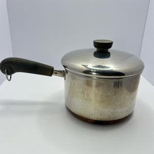 May include: A stainless steel saucepan with a polished finish, featuring a dark gray, textured handle with a small metal hanging loop at the end. The pot includes a matching stainless steel lid with a dark gray, circular knob on top.