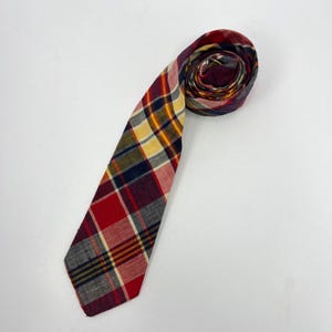 May include: A necktie featuring a multi-colored plaid pattern with intersecting lines in shades of red, navy blue, yellow, and cream. The fabric has a visible woven texture, and the tie is coiled at the top, displaying the layered fabric folds.