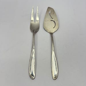 May include: A silver serving fork and pie server set. The fork has two tines and a long, curved handle. The pie server has a slotted blade and a matching handle. Both pieces are made of a shiny, reflective metal.