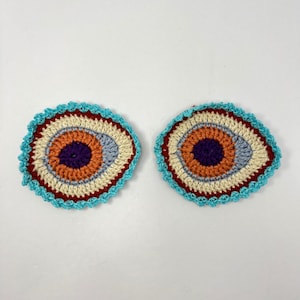 May include: Two handmade crochet coasters. Each coaster features a central purple circle, surrounded by orange, cream, and blue concentric rings, with a red border and a turquoise scalloped edge. The coasters are placed on a white surface.