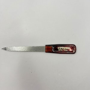 May include: A nail file with a silver abrasive surface and a dark brown handle. The handle has the text "REVLON SHAPING" in gold lettering. The file is designed for shaping and smoothing nails.