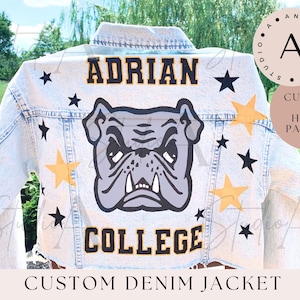 May include: A light blue denim jacket with a hand-painted design. The design features a bulldog, the words "Adrian College", and yellow stars. The jacket is customized and hand-painted.