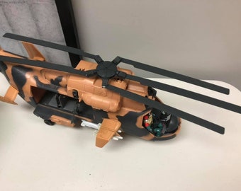 May include: A toy helicopter with a camouflage paint scheme. The helicopter has a black rotor system with four blades. The helicopter has a detailed cockpit with a pilot and a gunner.