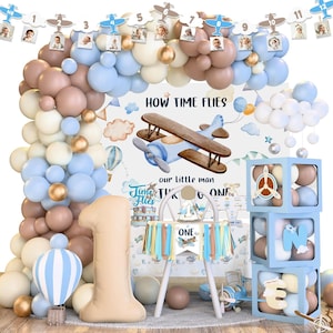 May include: A first birthday party decoration set with a blue and brown airplane theme. The set includes balloons, a high chair, a banner that says "ONE", and a large number "1" balloon. The backdrop features an airplane and the text "How Time Flies".