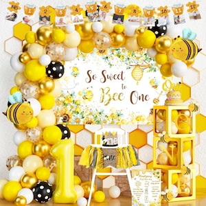 May include: A festive first birthday party decoration set with a bee theme. The display includes a balloon arch in yellow, gold, and white, a banner with photos, and a high chair with a "one" crown. The backdrop reads "So Sweet to Bee One."