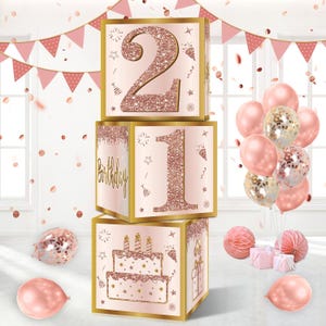 May include: A set of three pink and gold cardboard boxes with the numbers 21 and the word "Birthday" printed on them. The boxes are stacked on top of each other. There are pink and gold balloons in the background.