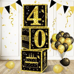 May include: Black and gold cardboard boxes with the numbers 40 on them. The boxes are stacked on top of each other. The top box has the number 4, the middle box has the number 0, and the bottom box has a birthday cake design. There are gold and black confetti and balloons in the background.