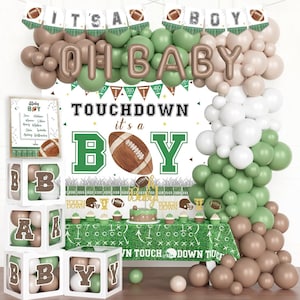May include: A football-themed baby shower decoration set. The decorations include a balloon arch in shades of green, brown, and white, with the words "OH BABY" and "IT'S A BOY". A table is covered with a football field tablecloth, and there are decorative boxes spelling out "BABY".