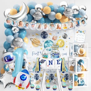 May include: A space-themed first birthday party decoration set. Includes a balloon arch with blue, silver, and gold balloons, a banner with baby photos, and a high chair with a space-themed cake. Also includes a rocket and astronaut balloon.