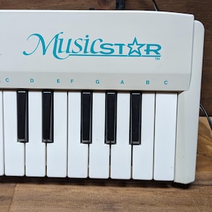 May include: A white electronic keyboard with black and white keys. The brand name MusicStar is printed in teal above the keys. Musical note letters C, D, E, F, G, A, B, and C are printed in black above the corresponding white keys.