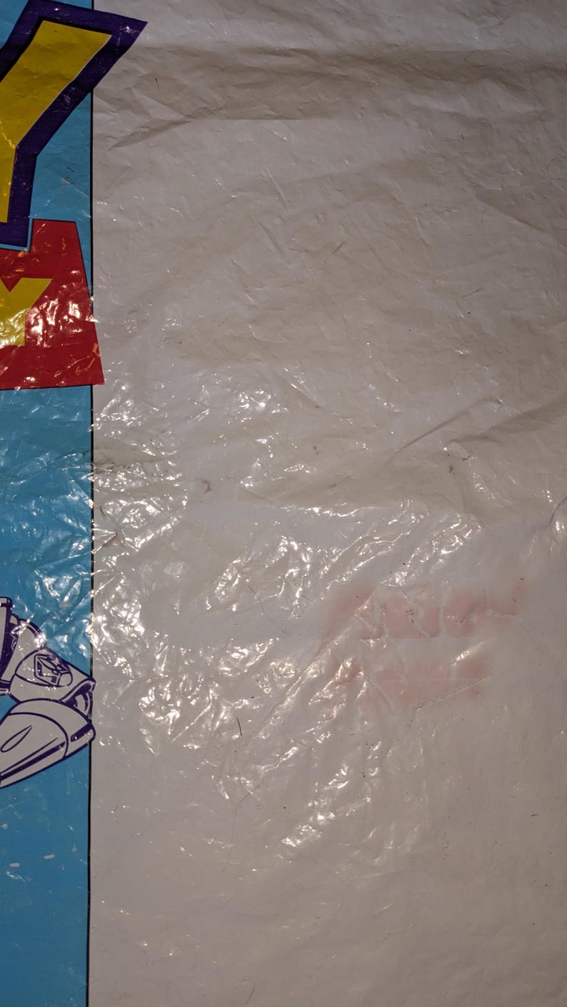 May include: A white plastic bag featuring a partial blue border on the left side. The visible graphic includes a red square with a yellow letter Y and a portion of a white and blue character illustration. The plastic surface is wrinkled and creased.