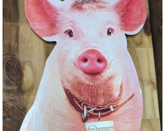 May include: A die-cut cardboard cutout of a pink pig with large ears and a snout. The pig wears a brown leather collar with a rectangular metal tag hanging from it. The tag features the word Babe printed in a script font with a gold accent.