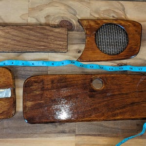May include: Four polished wooden components with a dark brown finish, including a rectangular block, a curved piece with a circular mesh insert, a small curved piece with a metal inlay, and a long panel with a hole, measured by a blue tape.