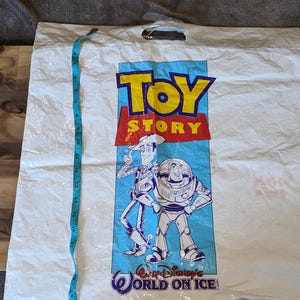 May include: A white plastic shopping bag featuring the Toy Story logo and characters Woody and Buzz Lightyear. The text Walt Disney's World on Ice is printed at the bottom. A blue measuring tape is placed vertically, showing a length of 45.7 cm.