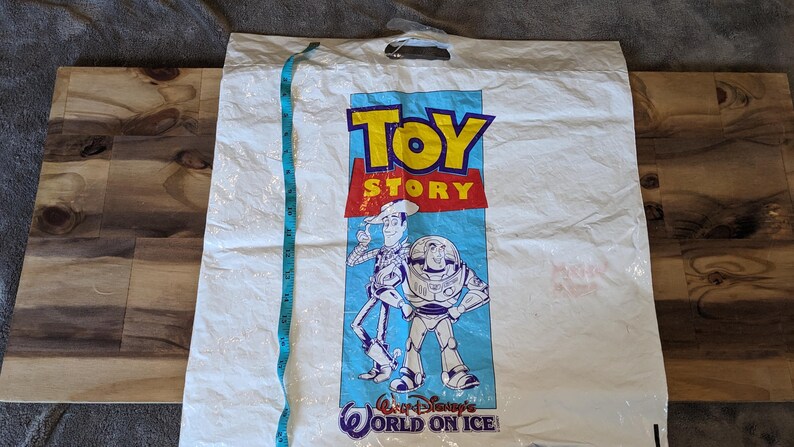 May include: A white plastic shopping bag featuring the Toy Story logo and characters Woody and Buzz Lightyear. The text Walt Disney's World on Ice is printed at the bottom. A blue measuring tape is placed vertically, showing a length of 45.7 cm.