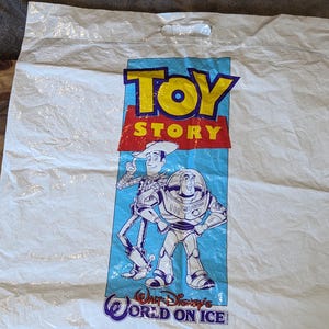 May include: A white plastic shopping bag featuring the Toy Story logo in yellow and red text. Below the logo, illustrations of Woody and Buzz Lightyear are printed on a light blue background, with the text Walt Disney's World on Ice at the bottom.