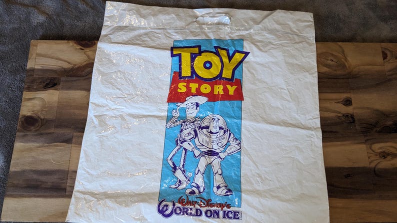 May include: A white plastic shopping bag featuring the Toy Story logo in yellow and red text. Below the logo, illustrations of Woody and Buzz Lightyear are printed on a light blue background, with the text Walt Disney's World on Ice at the bottom.