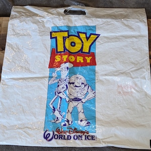 May include: A white plastic shopping bag featuring the Toy Story logo in yellow and red text. Below the logo, a graphic depicts the characters Woody and Buzz Lightyear. The text Walt Disney's WORLD ON ICE is printed in blue at the bottom.