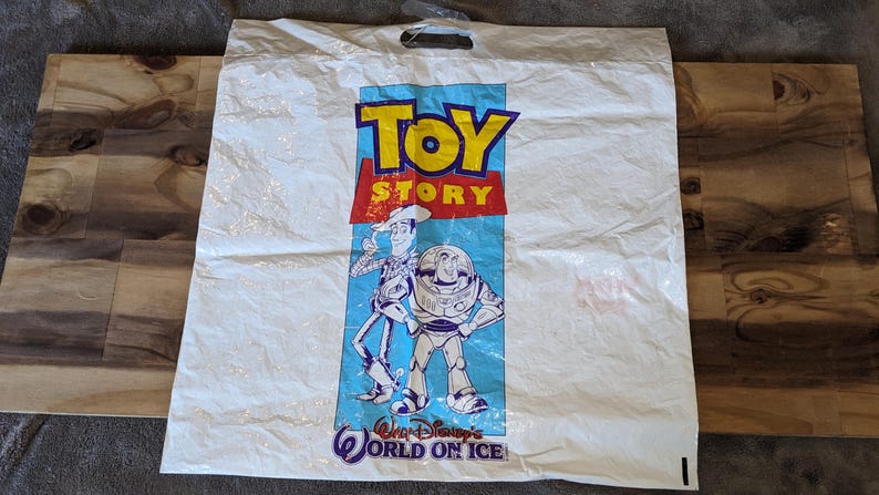 May include: A white plastic shopping bag featuring the Toy Story logo in yellow and red text. Below the logo, a graphic depicts the characters Woody and Buzz Lightyear. The text Walt Disney's WORLD ON ICE is printed in blue at the bottom.