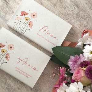 Peut inclure: Deux boîtes cadeaux blanches avec des motifs floraux et des lettres roses. La boîte du haut dit "Mama" avec le texte "Alles Liebe zum Muttertag" et la boîte du bas dit "Anne" avec le texte "ganz viel Glück zum allerersten Geburtstag" .