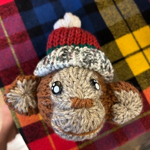 May include: A knitted monkey toy wearing a red and white knit hat with a white pom pom. The monkey has brown yarn for a nose and black button eyes.