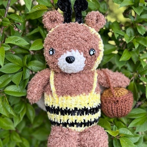 May include: A brown crocheted teddy bear wearing a yellow and black bee costume. The bear has black felt antennae and holds a brown crocheted basket.