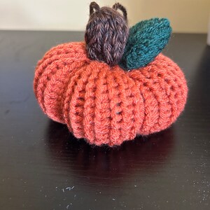 May include: A crocheted pumpkin with a brown stem and a green leaf. The pumpkin is orange and has a textured surface.