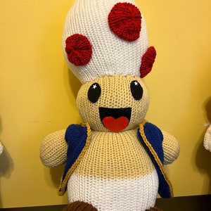 May include: A knitted plush toy shaped like a mushroom character with a white body, red spots, blue jacket, and brown shoes. The toy has a smiling face with black eyes and a red tongue.
