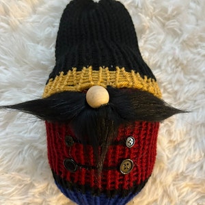 May include: A knitted gnome ornament with a black hat, gold trim, a wooden nose, a black moustache, and red and blue stripes. The gnome is wearing black buttons.