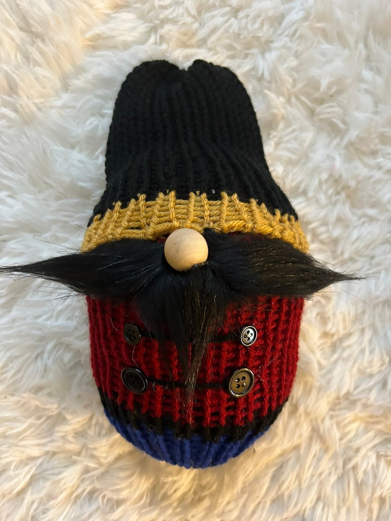 May include: A knitted gnome ornament with a black hat, gold trim, a wooden nose, a black moustache, and red and blue stripes. The gnome is wearing black buttons.