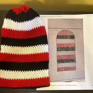 May include: A red, white, and black striped knitted beanie hat. The hat is laying flat on a table next to a printed beanie pattern worksheet.