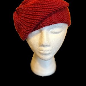 Könnte beinhalten: Eine rote Strickmütze mit einem einzigartigen gefalteten Design. Die Mütze ist aus weichem, strukturiertem Garn gefertigt und hat einen schlichten, klassischen Stil.