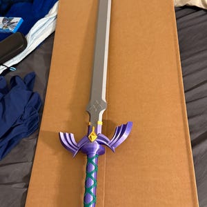 May include: A purple and silver fantasy sword with a green wrapped handle. The blade is silver, and the hilt is purple with gold accents. The handle is wrapped in a green crisscross pattern. The sword is resting on a brown cardboard surface.