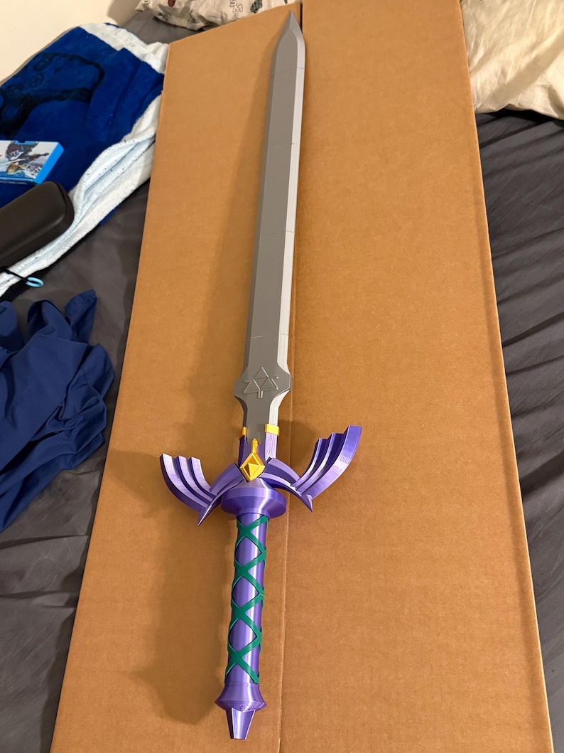 May include: A purple and silver fantasy sword with a green wrapped handle. The blade is silver, and the hilt is purple with gold accents. The handle is wrapped in a green crisscross pattern. The sword is resting on a brown cardboard surface.