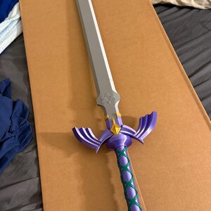 May include: A decorative sword with a silver blade and a purple hilt. The hilt features a green crisscross pattern and purple wings. A gold diamond shape is at the base of the blade. The sword is lying on a brown surface.